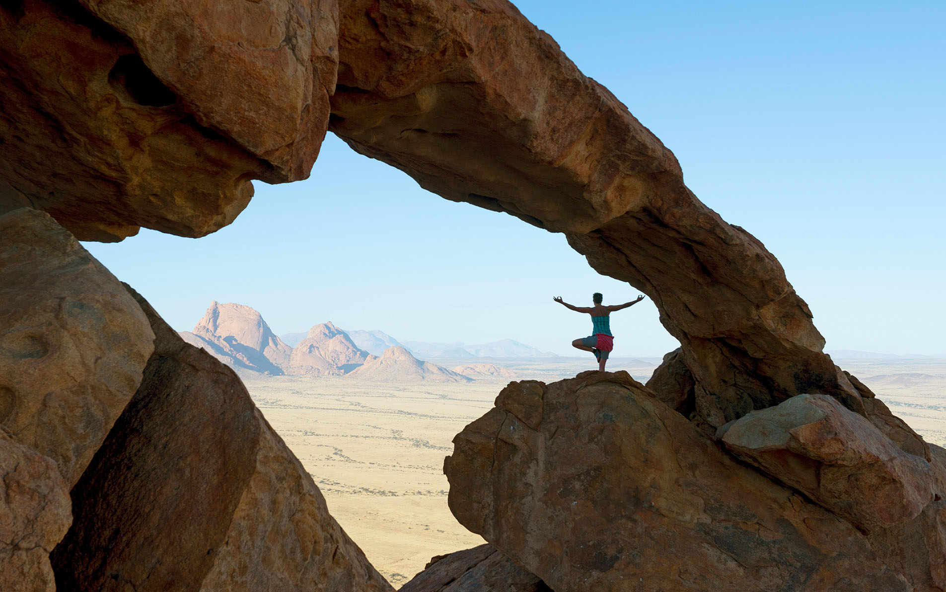 Woman doing yoga on rock formation in front of Spitzkoppe Mountain during bouldering expedition, Erongo region, Namibia