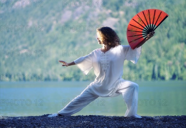 Young woman practicing Tai Chi Chuan with a red fan at mountain lake shore in the nature