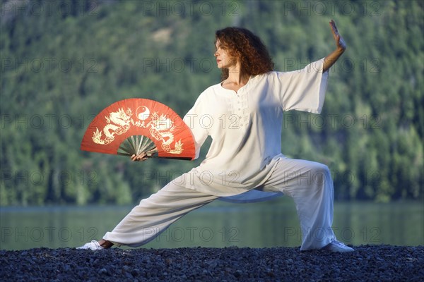 Young woman with a red hand fan practicing Taiji Pu Bu stance by the lake in the nature