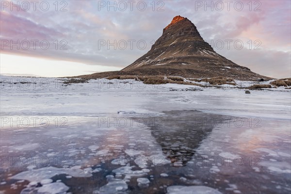 Mount Kirkjufell is reflected in the frozen lake