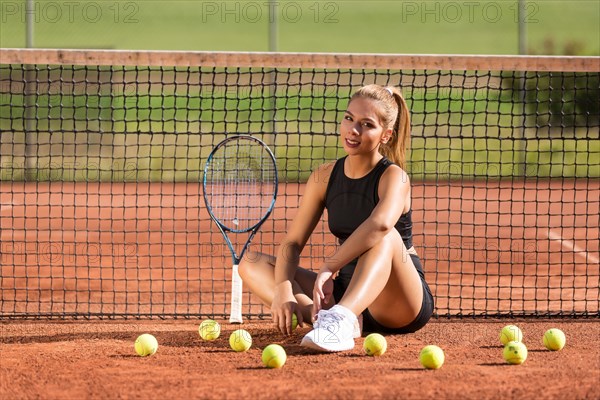 Young woman sitting in front of net with tennis racket and balls