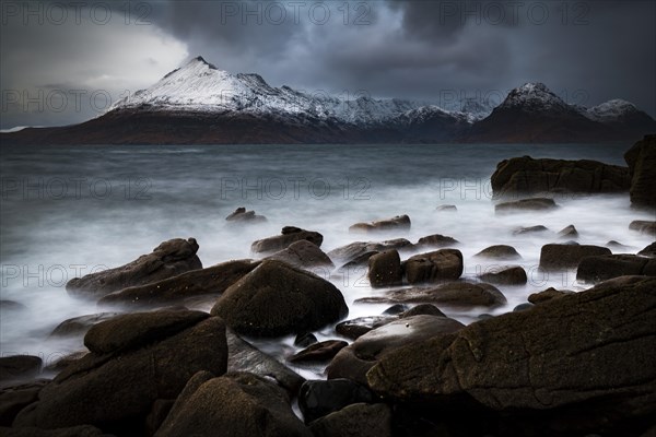 Big rocks in the water of the North Sea with snow-covered Cullin mountains in the background