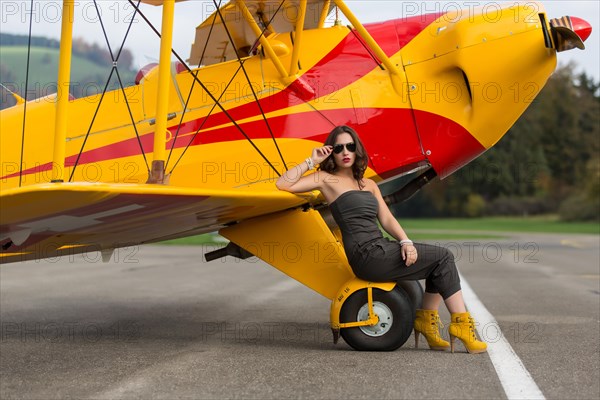 Young woman with sunglasses in overall and boots posing in front of double-decker airplane