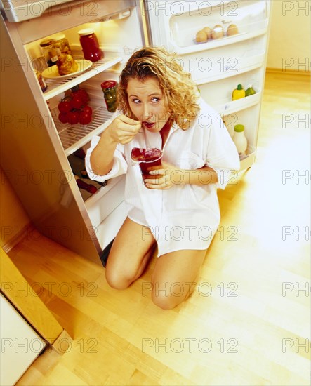 Woman kneeling in front of an open refrigerator in a nightshirt and eating red jelly