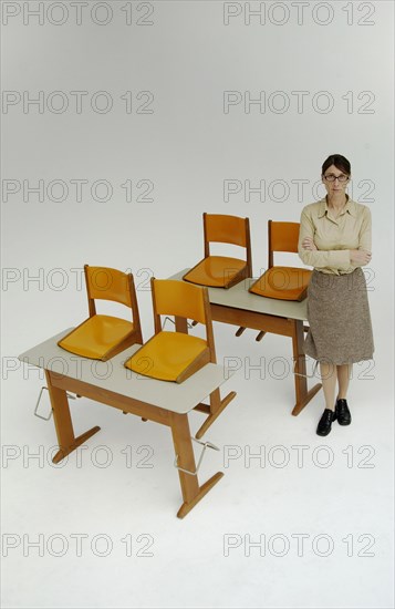 Strict female teacher standing next to old school desks with orange chairs
