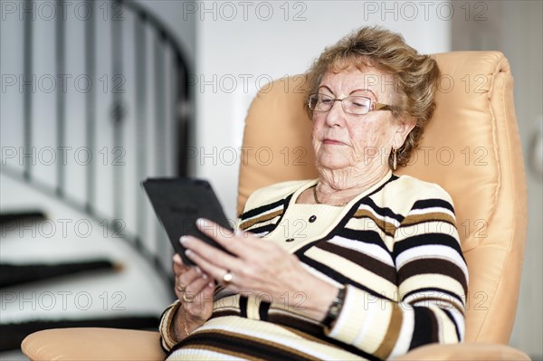 Elderly woman sitting in an armchair and reading an eBook