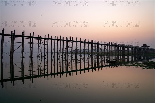 Buddhist monks walk across the U-leg bridge in red robes as the sun rises