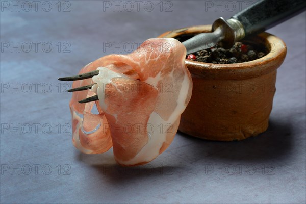 A slice of coppa on fork and clay pot with peppercorns