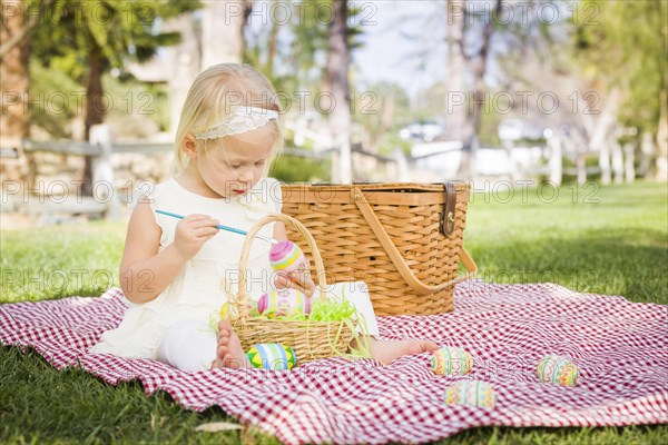 Cute baby girl enjoys coloring her easter eggs on picnic blanket in the grass