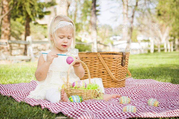 Cute baby girl enjoys coloring her easter eggs on picnic blanket in the grass