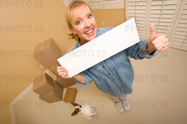 Excited woman with thumbs up and doggy holding blank sign near moving boxes in empty room taken with extreme wide angle lens