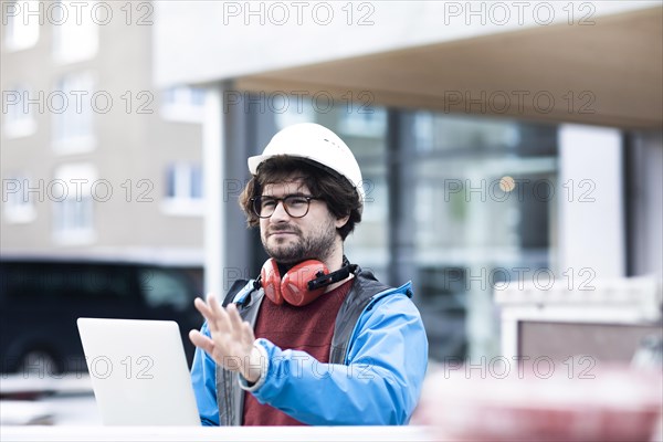 Young engineer with helmet and hearing protection checks outside work with laptop