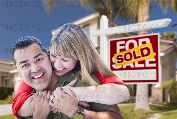 Happy hugging mixed-race couple in front of sold home for sale real estate sign and house