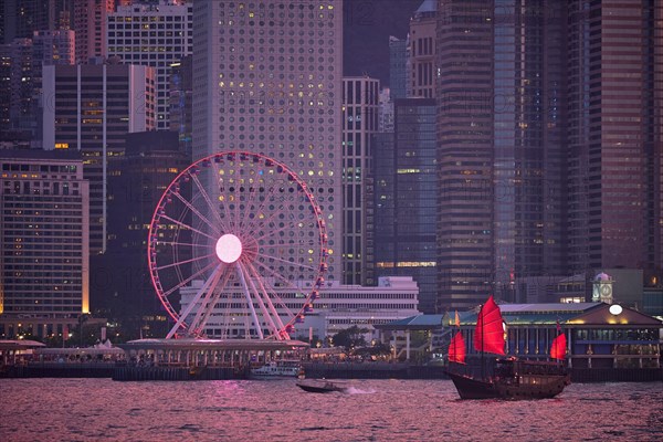 Tourist junk boat ferry with red sails and Hong Kong skyline cityscape downtown skyscrapers over Victoria Harbour in the evening. Hong Kong