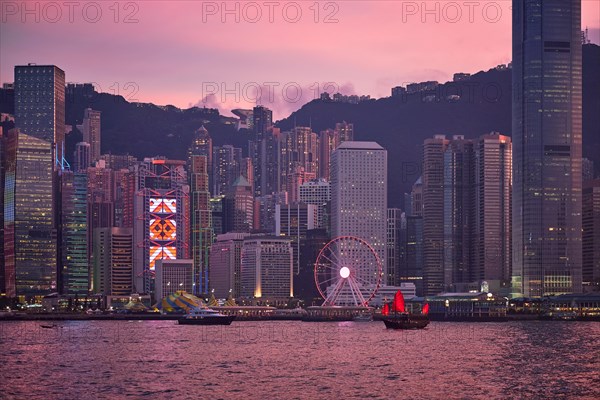 Tourist junk boat ferry with red sails and Hong Kong skyline cityscape downtown skyscrapers over Victoria Harbour in the evening. Hong Kong