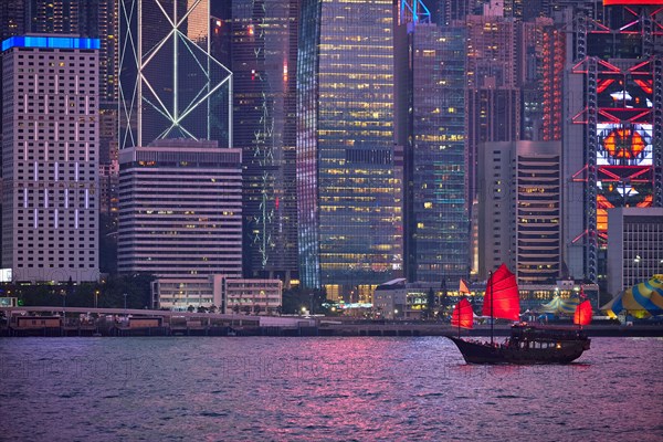 Tourist junk boat ferry with red sails and Hong Kong skyline cityscape downtown skyscrapers over Victoria Harbour in the evening. Hong Kong