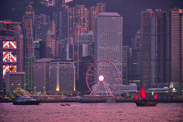 Tourist junk boat ferry with red sails and Hong Kong skyline cityscape downtown skyscrapers over Victoria Harbour in the evening. Hong Kong