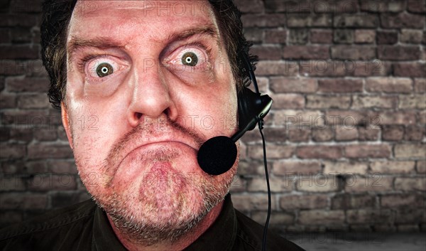 Grumpy businessman with frowning face wearing phone headset in office with brick background