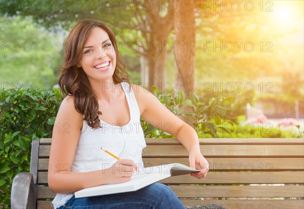 Attractive young adult female student on bench outdoors with books and pencil