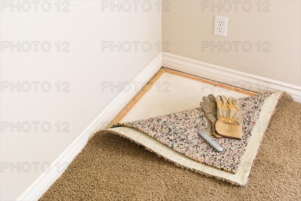 Construction gloves and utility knife on pulled back carpet and underlay in room