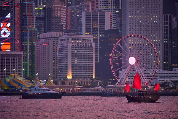 Tourist junk boat ferry with red sails and Hong Kong skyline cityscape downtown skyscrapers over Victoria Harbour in the evening. Hong Kong