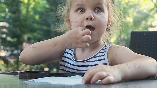 Little girl tastes salt. Close-up portrait of blonde girl takes salt from napkin with her finger and tastes it while sitting in street cafe on the park. Odessa