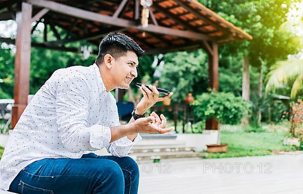 Man sitting sending a voice message with his phone and gesturing with his hands. Handsome man sitting outside sending a voice message with his cell phone
