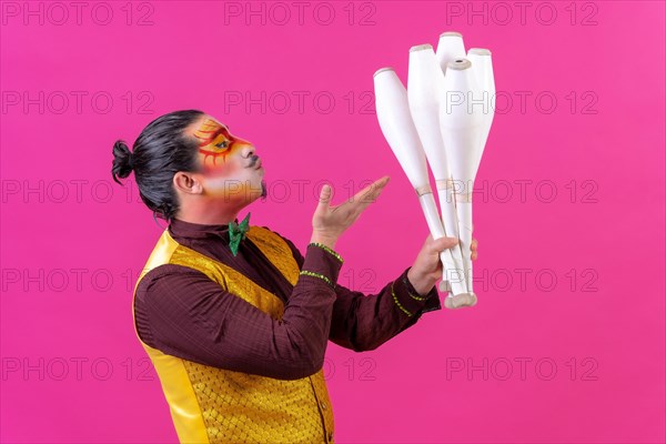 Juggler in a vest and with painted face juggling maces on a pink background