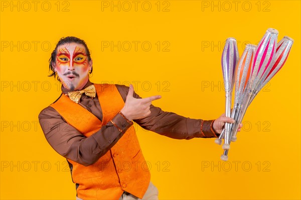 Portrait of a happy juggler man in make up vest juggling with maces on a yellow background