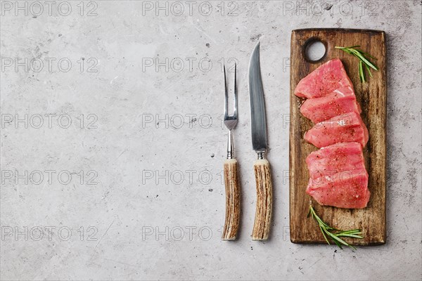 Top view of pork fillet sliced into pieces on wooden cutting board next to knife and fork for grill