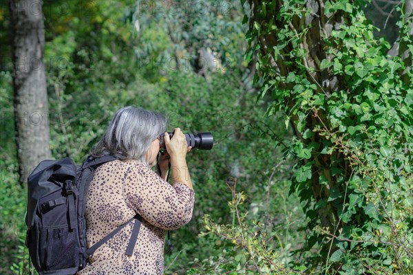 White-haired female photographer in profile and backpack view taking pictures of a ivy