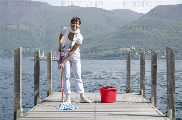 Woman scrubs the Pier over the alpine lake with cleaning equipment and mountain in background in Ticino