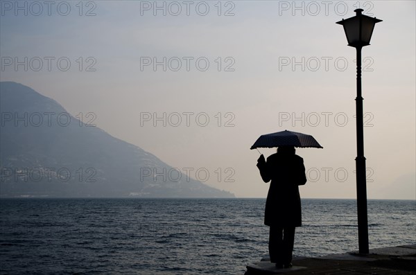 Woman Standing on the Waterfront with an Umbrella close to a Street Lamp