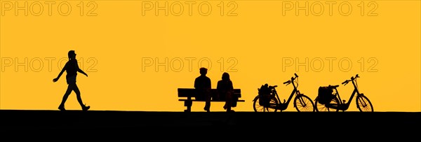 Girl walking past couple of elderly cyclists resting on bench next to their two bicycles silhouetted against yellow sunset sky in summer