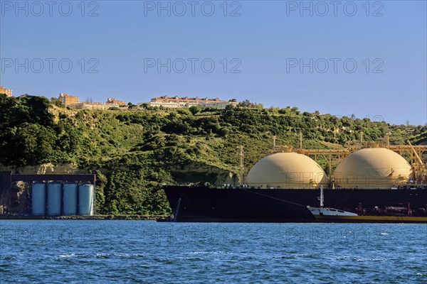 LNG or liquified natural gas tanker moored by industrial pier and green hill on sunny day. Alternative gas supply