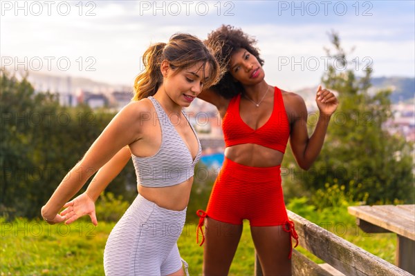 Caucasian blonde girl and dark-skinned girl with afro hair doing stretching before starting sports in the park. Healthy life