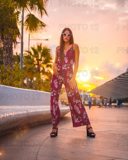 Sunset with a young brunette in a maroon floral dress and modern white sunglasses enjoying the summer in the golden hour