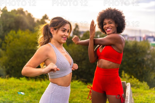 Caucasian blonde girl and dark-skinned girl with afro hair doing stretching before starting sports in the park. Healthy life