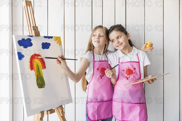 Happy girl holding palette hand looking her friend painting canvas with brush
