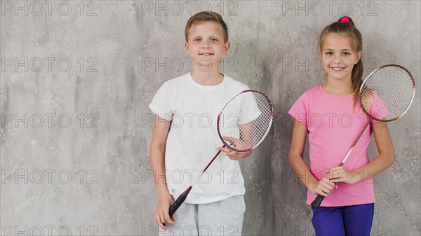 Portrait smiling boy girl holding racket hands against concrete wall