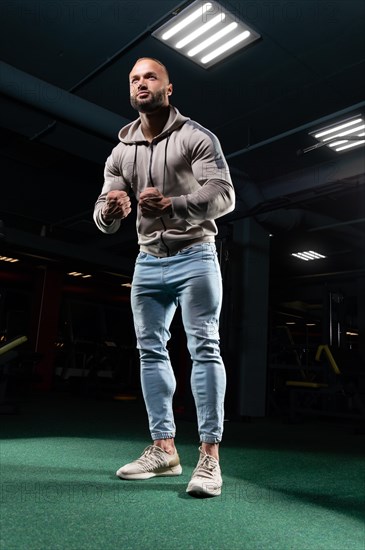 Muscular man poses in the gym in jeans and a sweatshirt. Sports