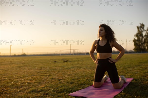Young curly athletic girl in sportswear doing yoga on a yoga mat outdoors on the grass during sunset
