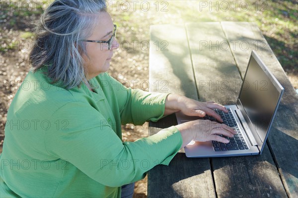 Mature woman with white hair and glasses working on her computer in the sunlit countryside