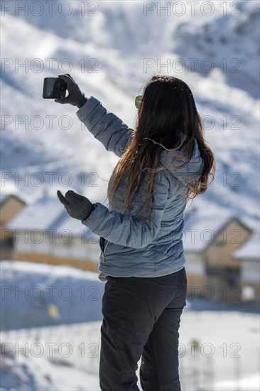 A woman in casual winter clothing takes a photo with her smartphone in a snowy mountainous area