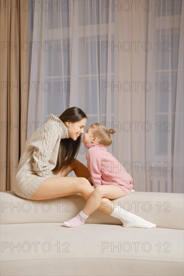 A mother and daughter sit on sofa in front of each other and touching each other's noses in a well-lit living room