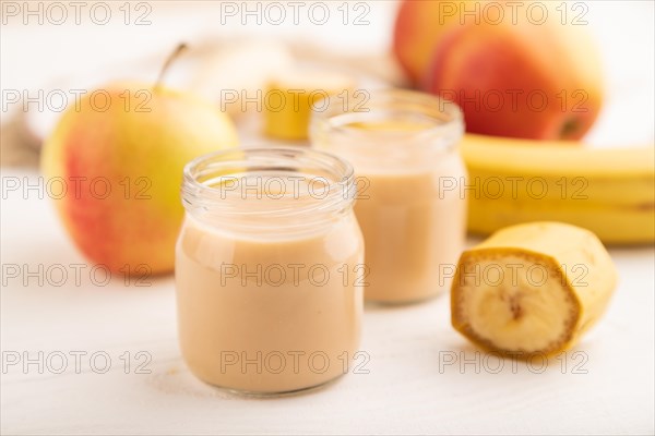 Baby puree with fruits mix, apple, banana infant formula in glass jar on white wooden background. Side view, close up, selective focus, artificial feeding concept