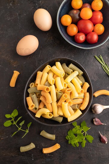 Rigatoni colored raw pasta with tomato, eggs, spices, herbs on black concrete background. Top view, flat lay