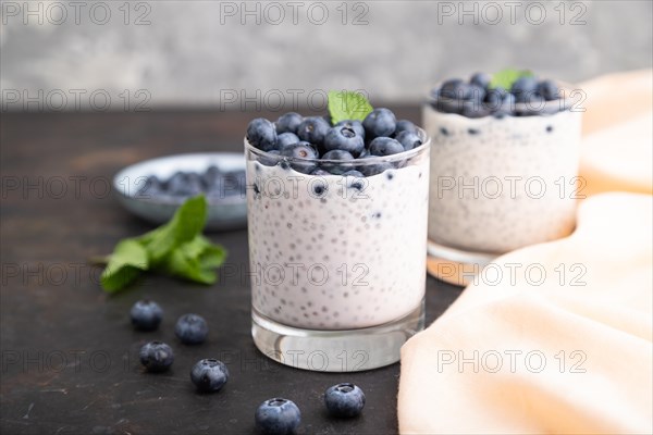 Yogurt with blueberry and chia in glass on black concrete background and orange linen textile. Side view, close up