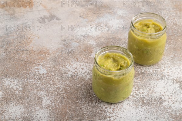 Baby puree with vegetable mix, broccoli, tomatoes, cucumber, avocado infant formula in glass jar on brown concrete background. Side view, copy space, artificial feeding concept
