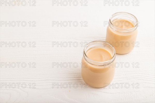 Baby puree with fruits mix, apple, banana infant formula in glass jar on white wooden background. Side view, copy space, artificial feeding concept